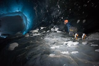Un uomo visto dal basso si aggrappa alla parete di ghiaccio, alzando lo sguardo verso il soffitto ghiacciato