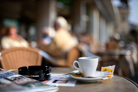 Una tazzina di caffè, una bustina di zucchero e un giornale su un tavolino all aperto. Avventori del bar sfocati.
