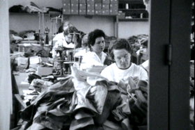 Donne sedute alla macchina da cucire circondate da scampoli di tessuto; sul fondo confezioni accatastate