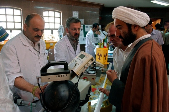 Tecnici in camice bianco mostrano apparecchiature a un ispettore con turbante. Accanto, simbolo di radioattività