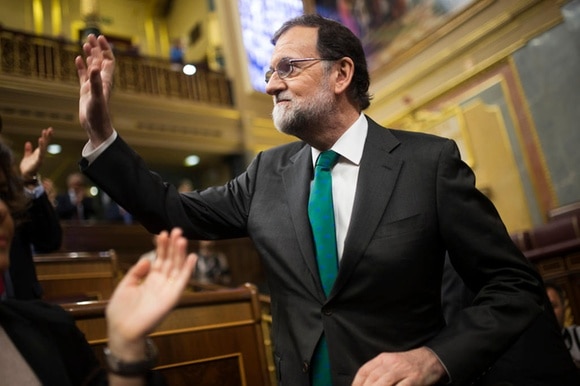 Primo piano di mariano Rajoy che alza la mano in segno di saluto; sullo sfondo, scranni del parlamento sfocati