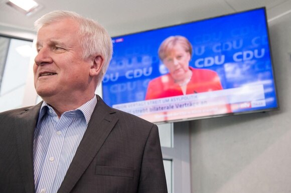 il ministro dell interno tedesco seehofer con alle spalle una televisione con l immagine di Angela Merkel