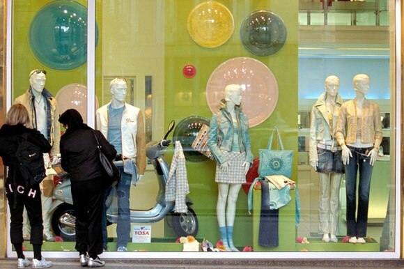 Vetrina di un negozio di vestiti; due donne di schiena, sulla sinistra, guardano un capo nel dettaglio