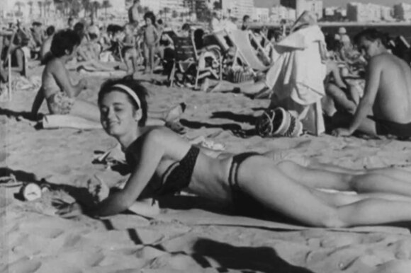 Donna distesa al sole, sulla spiaggia; un uomo dietro di lei la guarda; sul fondo lungomare con palazzi