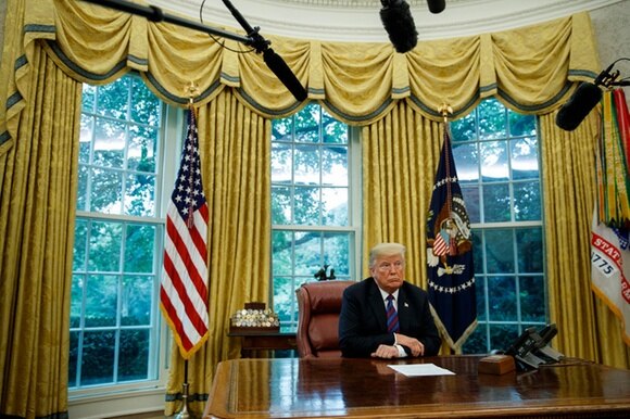 Trump seduto alla scrivania dello Studio ovale con dei foglia davanti a sé; bandiere, tende e microfoni a giraffa tutt intorno