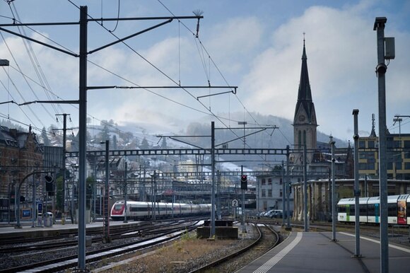Immagine di stazione ferroviaria; in primo piano banchine e binari; due convogli in arrivo; cittadina sul fondo