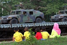 Bambini seduti sul ciglio della strada guardano passare veicoli blindati con militari armati di mitra.