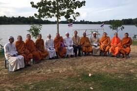 Un gruppo di uomini, buddhisti e musulmani, è seduto a semicerchio su un prato con il mare alle spalle.