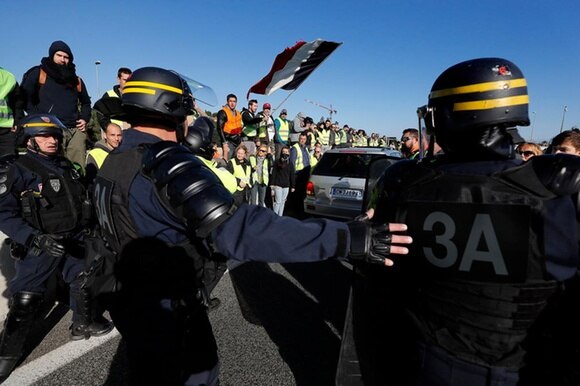 Auto si fa strada in una folla di persone che indossano mantellina catarifrangente gialla; poliziotti con casco e scudo intorno