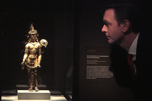 Uomo di profilo e in penombra osserva una scultura buddista, ben illuminata, in una teca