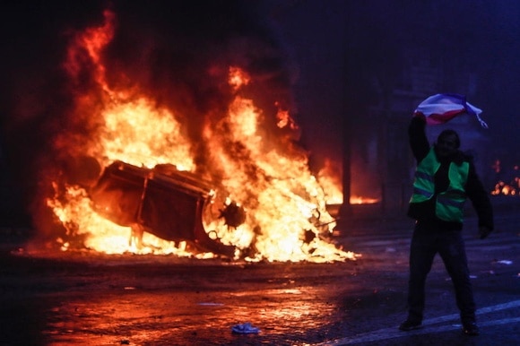UN uomo con il gilet giallo con la bandiera francese mentre dietro un auto sta bruciando