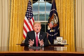 Donald Trump seduto alla scrivania parla gesticolando; dietro, badiera americana e tende/finestra, accanto, foto ritratto