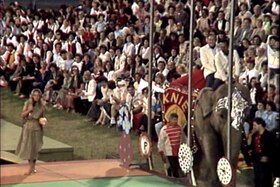 Due uomini in abito bianco in groppa a un elefante; dietro, platea; accanto, presentatrice con microfono