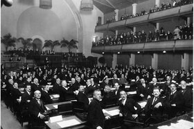 Una sala da assemblea, con banchi e balconate, colma di uomini in abito formale