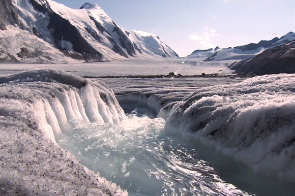 Veduta di un ghiacciaio (non identificato), con montagne innevate ai lati e acqua che scorre in primo piano