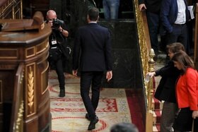 Uomo ritratto di schiena mentre lascia un aula parlamentare (si distinguono un pulpito e banchi); un fotografo lo ritrae
