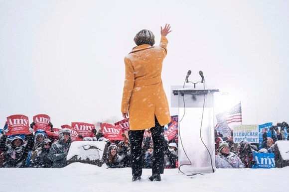 Una donna, di schiena, accanto a un pulpito con microfoni saluta folla sotto la neve che regge cartelli con scritto Amy