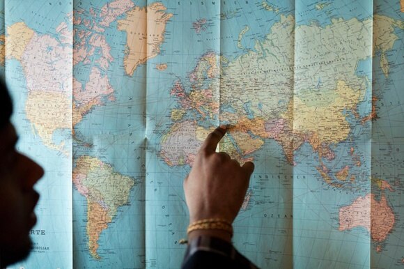 Mano indica un percorso su una cartina del mondo. Sulla sx, si intravvede di profilo il volto della persona