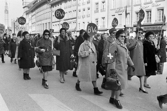 Immagine in bianco e nero di donne in cappotto che sfilano reggendo cartelli circolari con la scritta BV4