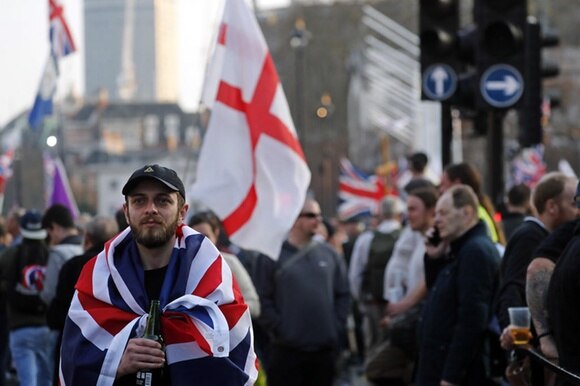 In primo piano, uomo avvolto in una bandiera britannica guarda sconsolato; dietro, sfocata, folla di manifestanti