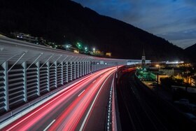 Tratto autostradale con ripari fonici e traliccio con cartello di limite di velocità dinamico, ripreso di notte (scie rosse)