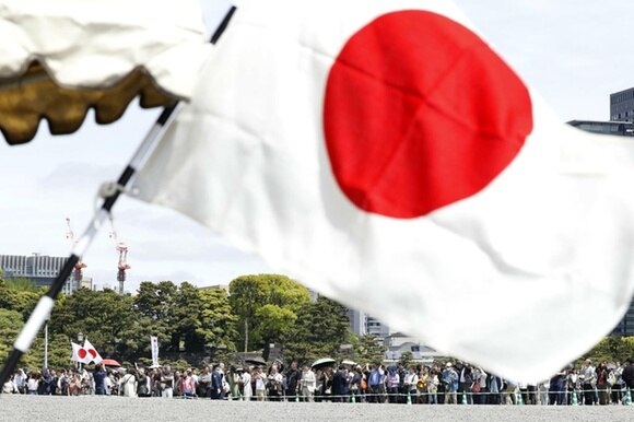 Primissimo piano di una bandiera giapponese. In lontananza, adunata di persone ai margini di un parco