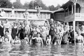 Gruppo di uomini e donne in acqua sul litorale di un lago; indossano costumi d altri tempi; costruzione in legno sul fondo