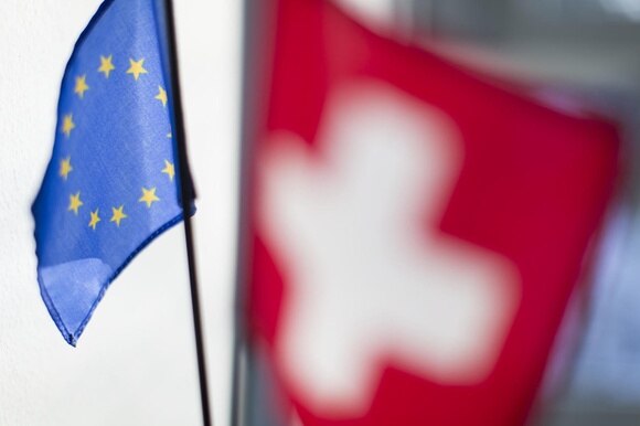Primo piano di una bandierina UE (a fuoco) con sullo sfondo una bandiera svizzera) sfocata