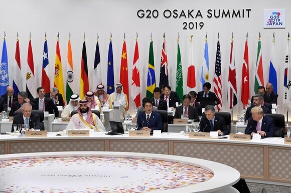 Ampio banco leggermente curvo con Abe al centro e altri politici tutt attorno; sul fondo bandiere e logo del G20