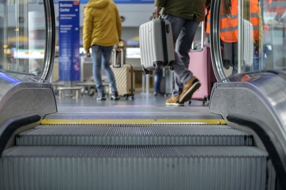 Parte finale di una scala mobile in ambiente aeroportuale; due persone col trolley e cartelli indicatori