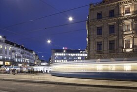 Vista di una piazza al crepuscolo con edificio con finestre illuminate in fondo e tram che sfreccia in primo piano.