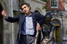 Placido Domingo in camicia e giacca blu posa per i fotografi accanto a una scultura con un espressione ironica