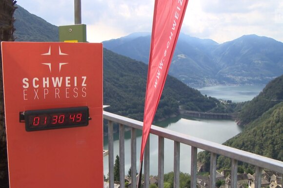 Vista dall alto di un bacino lacustre circondato da montagne, con totem con scritta Schweiz express e cronometro