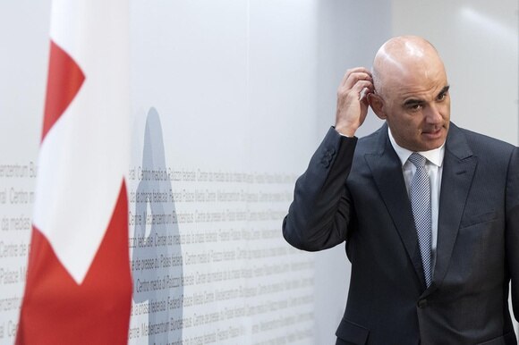 Alain Berset a mezzobusto, sulla dx, si gratticchia la testa; sulla sx, bandiera svizzera e sfondo della sala stampa