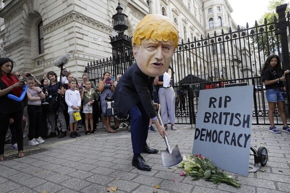 uomo travestito da Boris JOhnson con una pala davanti a un cancello e un cartello