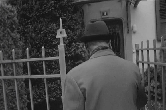 Uomo con cappotto e cappello, di schiena, varca il cancelletto di una palazzina numero civico 11