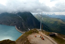 Veduta aerea di una diga; su un promontorio vicino un generatore eolico in fase di installazione
