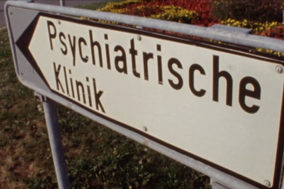 Primissimo piano di un cartello con scritto Psychiatrische Klinik, installato in una aiuola coltivata a fiori