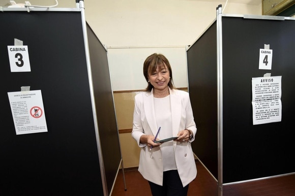 Donna in pantaloni nero e blazer bianco fa capolino tra due cabine elettorali con in mano delle schede e una penna