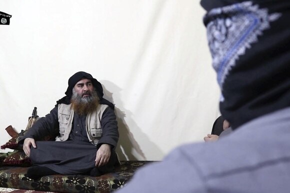 Uomo con abiti arabi e giberna da combattimento seduto a gambe incrociate con accanto un kalashnikov
