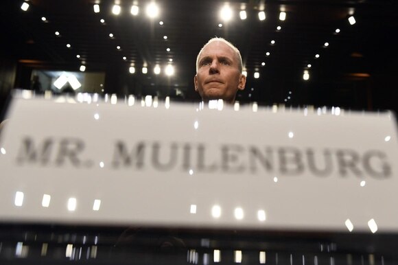 Primo piano di cartello, sfocato, con il nome MR. MUILENBURG; dietro, a fuoco, il volto dello stesso sovrastato da luci