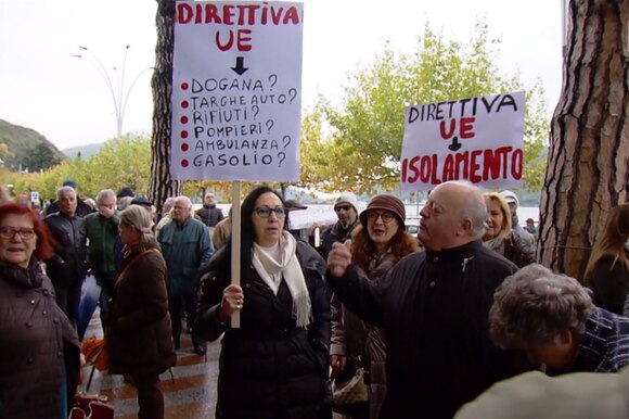 Gruppo di persone manifesta con cartelli vari; uno riporta il quesito riportato nella didascalia