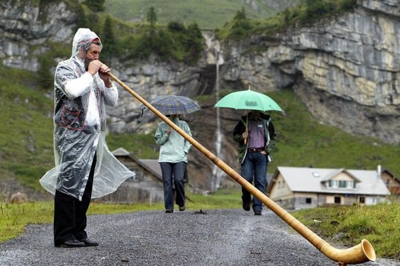 Un uomo suona il corno delle Alpi mentre piove, coperto da una mantellina, in ambiente montano