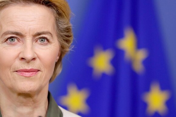 Primissimo piano di Usrula Von Der Leyen; si intuisce una bandiera europea dietro di lei