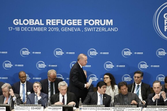 Gruppo di uomini (una sola donna) seduti a un tavolo sotto la scritta Global Refugee Forum ; Erdogan in piedi con fogli in mano