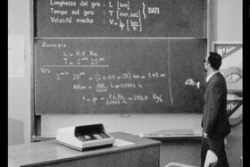 Immagine b/n di un maestro che spiega formule matematiche alla lavagna.