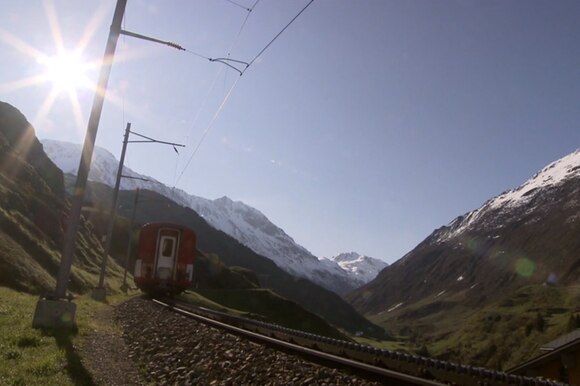 Immagine controsole di una vallata alpina percorsa da binari; un treno si allontana