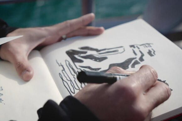 Primo piano di uno sketchbook, una mano lo tiene fermo, l altra stilizza delle montagne a pennarello nero.