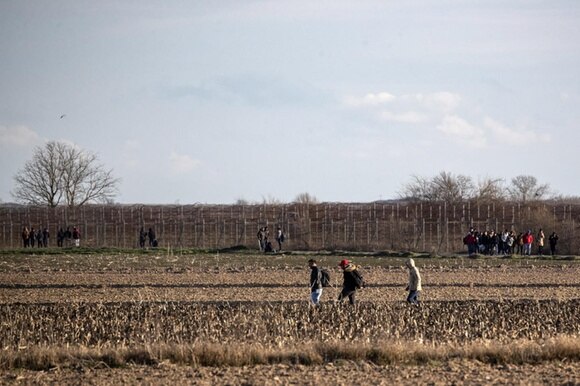 Campi coltivati; si vedono gruppi di persone che camminano e, in lontananza, una fitta rete metallica