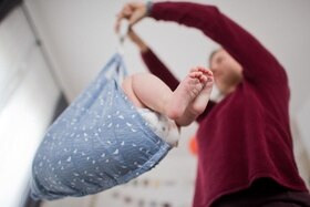 Signora vista dal basso (viso sfocato) pesa un bambino con una bilancia per neonati; spuntano le gambe del bebé